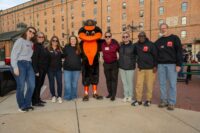 The Baltimore Station staff and WBFF Fox45 meteorologist and event EMCEE Justin Chambers (fourth from right) pose with the Oriole Bird at the Stars, Stripes and Chow chili cook-off fundraising event. (The Baltimore Station)